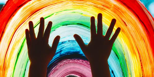 childs hands pressed against a window which has been painted with a rainbow using colours, red, orange, yellow, green, blue, purple.