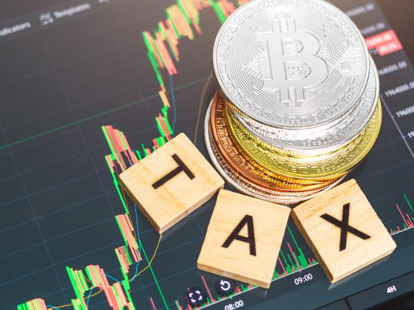 a tablet showing a cryptocurrency graph, a stack of Cryptocurrency, and the word 'TAX' arranged around the stack of coins with wooden scrabble tiles