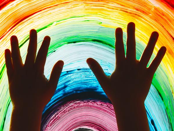 childs hands pressed against a window which has been painted with a rainbow using colours, red, orange, yellow, green, blue, purple.