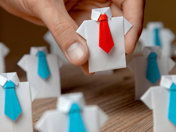 a desk of little white origami shirts wearing coloured tie's, all of which have blue ties except for the one in the persons hand which is wearing a red tie.