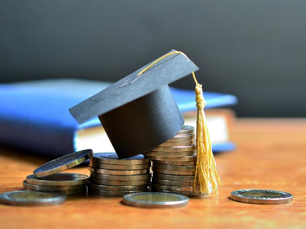 a pile of coins on a wooden table, in the background sits a blue book. On top of the coin pile is a tiny black graduation cap
