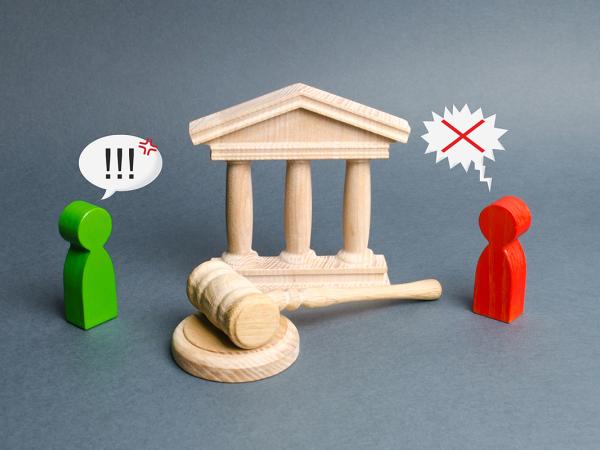 a wooden made figure of a courthouse, a wooden Gavel and 2 small wooden people with speech bubbles above their heads, the green person's speech bubble shows '!!!' the red one's speech bubble is jagged edged and shows 'X'