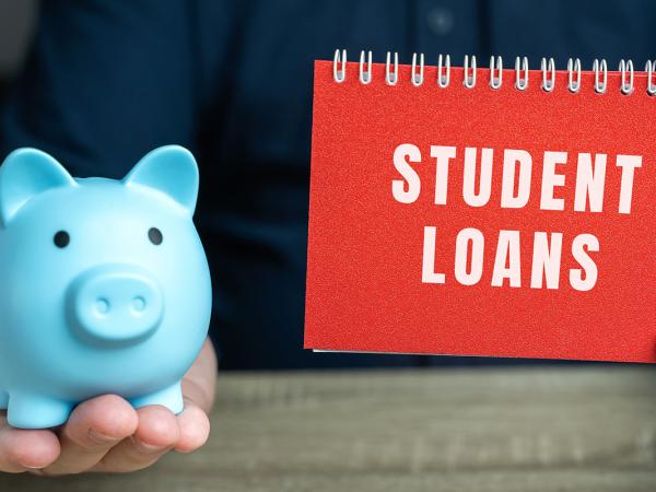 a person holding a blue piggy bank and a book named 'STUDENT LOANS' 