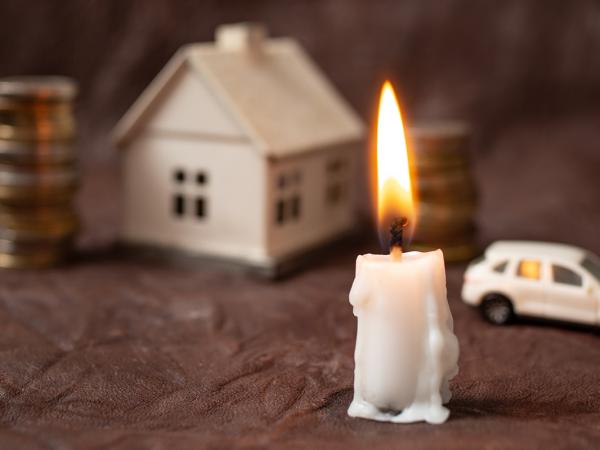 a lit candle, in the background a stack of coins, a house, and a car can be seen. 