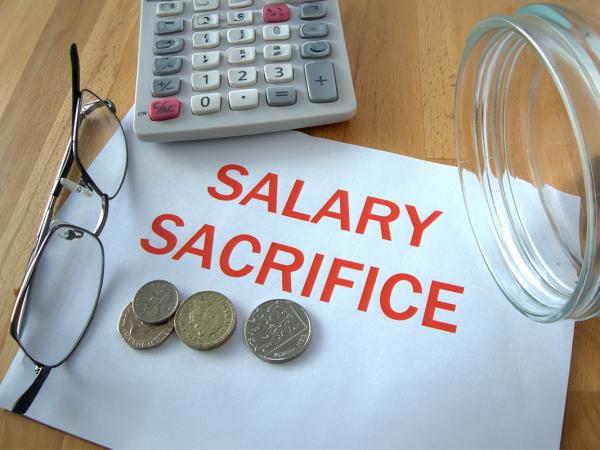 a sheet of white paper with the words 'SALARY SACRIFICE' typed on them in red ink. next to this is a pair of glasses, a calculator an a jar of money tipped on its side, some of the money has made it's way onto the paper. 