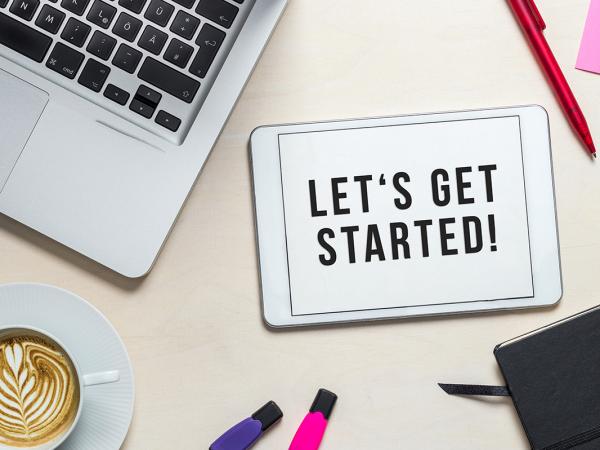 a desk with a laptop, pen, coffee, diary, highlighters and a tablet with the words 'LETS GET STARTED' on the screen. 