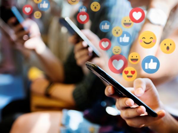 people looking at social media on their mobile phones, surrounded by social media reaction icons such as thumbs up, heart, smiley face. 