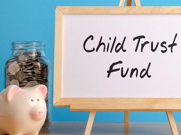 a wooden easel with the words 'CHILD TRUST FUND' written in black pen, next to this a piggy bank and a jar of coins can be seen against a blue background.