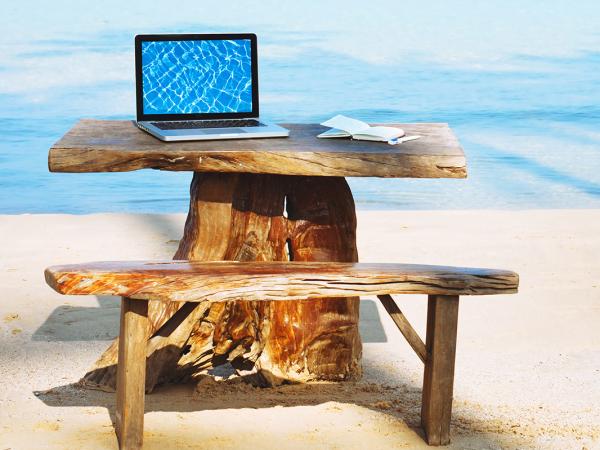 a wooden desk and seat set up looking over the sea abroad, on the desk sits a laptop.