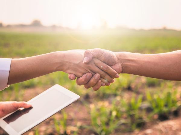 2 people shaking hands, one is holding a tablet in their other hand. The background is of countryside.