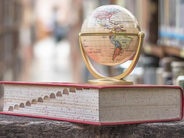 a large textbook with a globe of the world sat on top. background shows a library with many shelves of books.