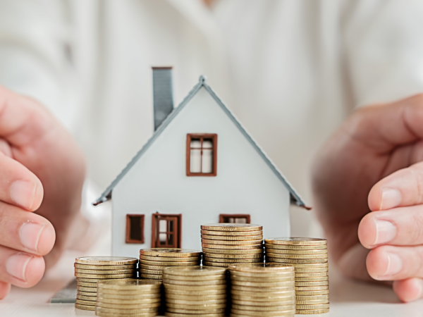 A small model house with stacks of coins outside, a person is cupping their hands on either side of the house. 