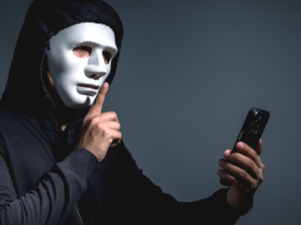 A person wearing all black with a white mask making the 'SSHHH' sign with their hand, in the other hand they are holding a phone. 