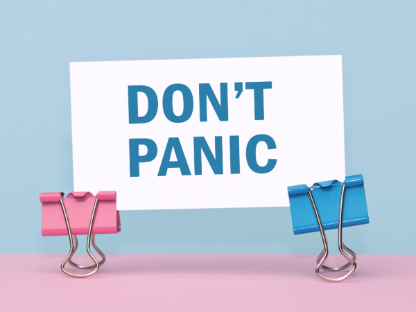 a piece of white paper with the words 'DON'T PANIC' printed in blue text, the paper is held up by 2 coloured foldback paper grips against a pink and blue background. 