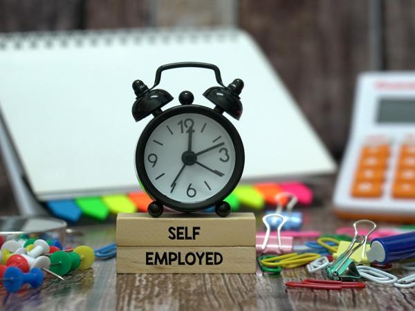 A desk scattered with various stationary, a clock and wooden blocks with the words 'SELF EPLOYED' printed on them in black text. 