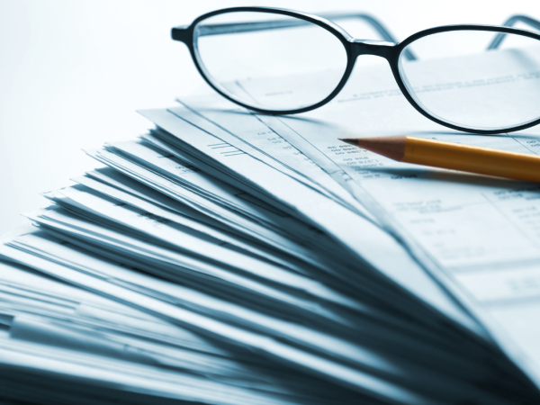 A large stack of papers with a pair of glasses and a pencil on top. 