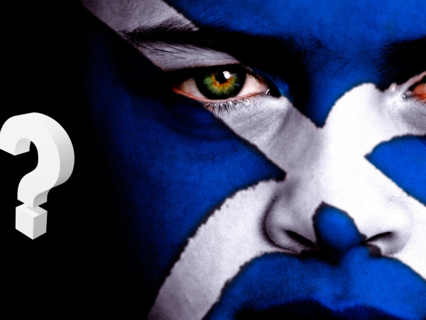 A person with their face painted as the Scottish flag against a black background, in the background a white question mark can be seen. 