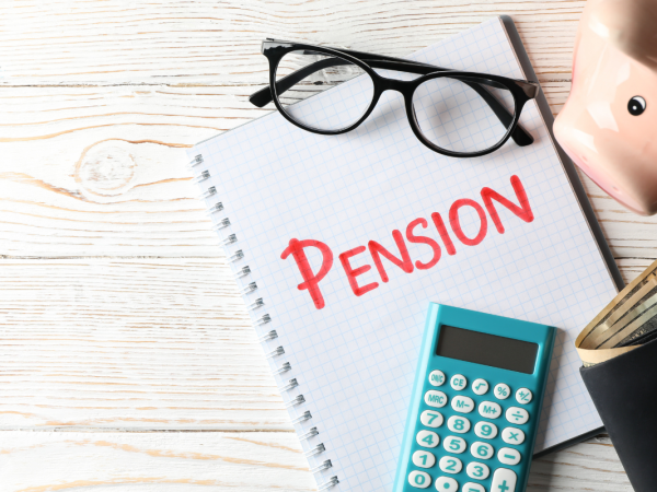 A desk with a piggy bank, a wallet,pair of glasses, a calculator and a notepad with the word 'PENSION' written on the front in red marker pen.