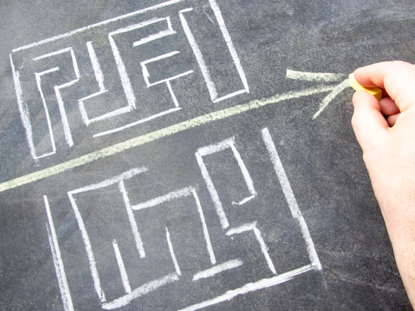 two hand drawn mazes on a chalkboard, and a person drawing a straight arrow through the gap between the two. 