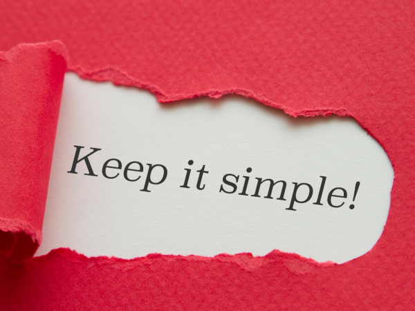 Red paper with a tear in the centre, through the tear the words 'KEEP IT SIMPLE!' can be seen typed onto a white paper.