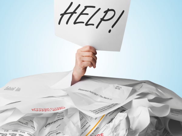 A person buried beneath a stack of overdue papers, a hand can be seen sticking out holding up a piece of paper with the word 'HELP!' written on it in black ink. 