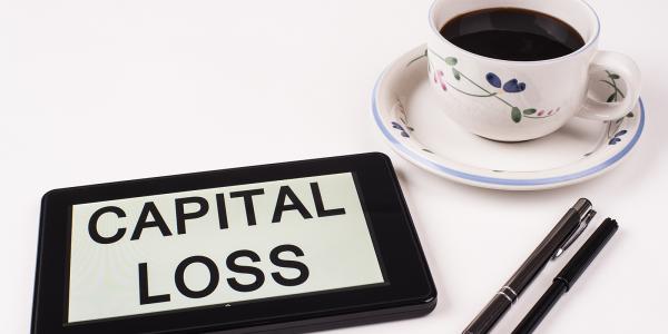 a tablet with the words 'CAPITAL LOSS' typed on the screen, two pens and a cup of black coffee. 