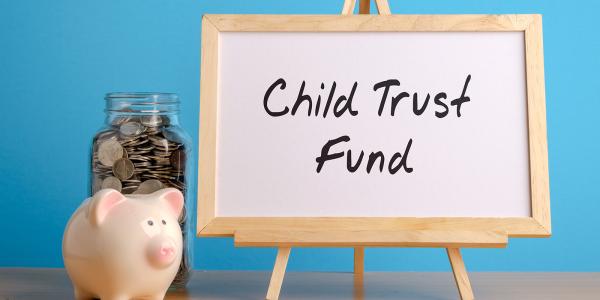 a wooden easel with the words 'CHILD TRUST FUND' written in black pen, next to this a piggy bank and a jar of coins can be seen against a blue background.