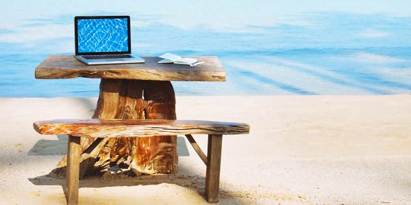 a wooden desk and seat set up looking over the sea abroad, on the desk sits a laptop.