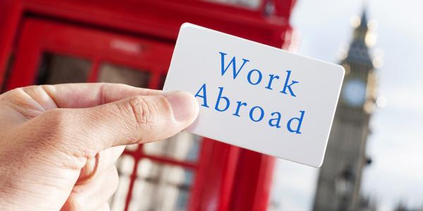 a person holding up a card that says 'WORK ABROAD' in blue text. The background shows the picture was taken in London as Big Ben and a red telephone box can be seen. 