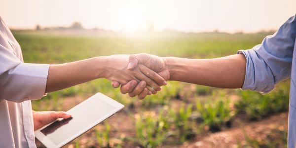 2 people shaking hands, one is holding a tablet in their other hand. The background is of countryside.