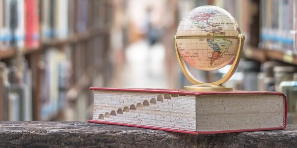 a large textbook with a globe of the world sat on top. background shows a library with many shelves of books.