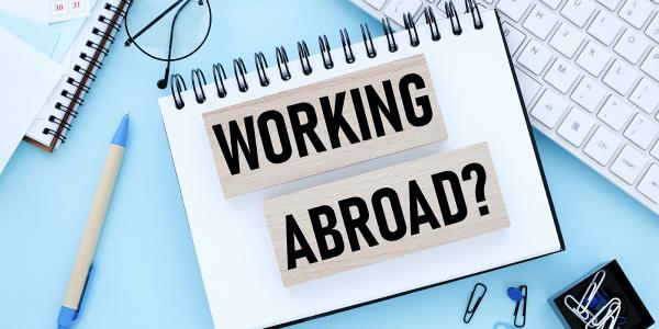 Two wooden blocks with the words 'WORKING ABROAD' showing on them in black text  sat on top of a notebook on a persons desk. Scattered around the notebook various stationary can be seen and a computer keyboard.