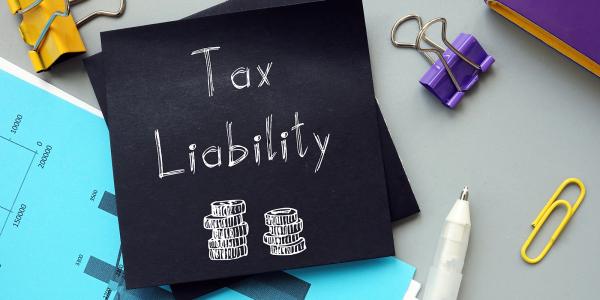 Black sheet of paper with the words 'TAX LIABILITY' written in white with an illustration of 2 piles of coins, various stationary surrounds the piece of black paper. 