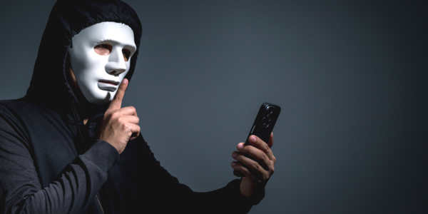 A person wearing all black with a white mask making the 'SSHHH' sign with their hand, in the other hand they are holding a phone. 