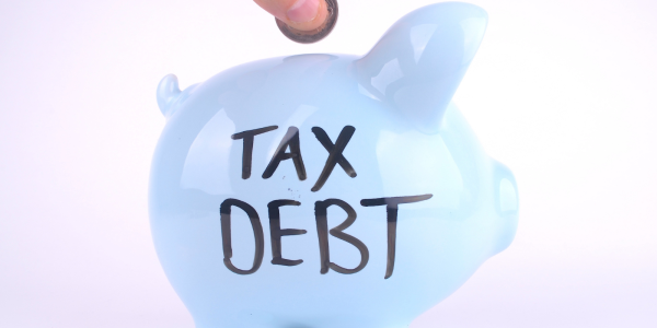 A person putting coins into a light coloured piggy bank with the words 'TAX DEBT' written in black ink. 