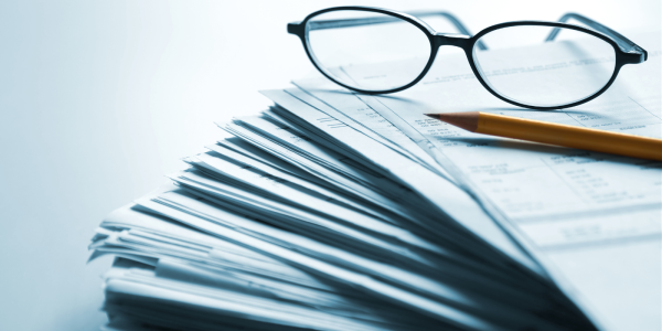 A large stack of papers with a pair of glasses and a pencil on top. 