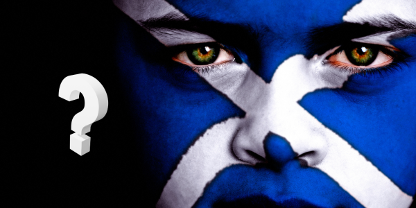 A person with their face painted as the Scottish flag against a black background, in the background a white question mark can be seen. 