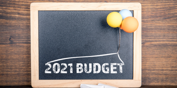 A chalkboard with the words '2021 BUDGET' printed on it, a small bunch o balloons are attached to the chalkboard.