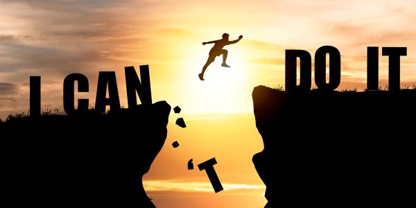 A person jumping from one cliff edge to another, the words 'I CAN DO IT' can be seen across the cliffs. The jump has knocked the 'T' from 'CAN'T'.