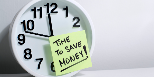 A wall clock with a post it note stuck to the front, the note reads 'TIME TO SAVE MONEY!'