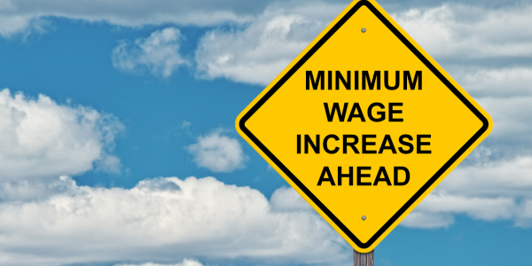 A blue sky with a scattering of white clouds and a yellow road sign with the words 'MINIMUM WAGE INCREASE AHEAD' in black text.