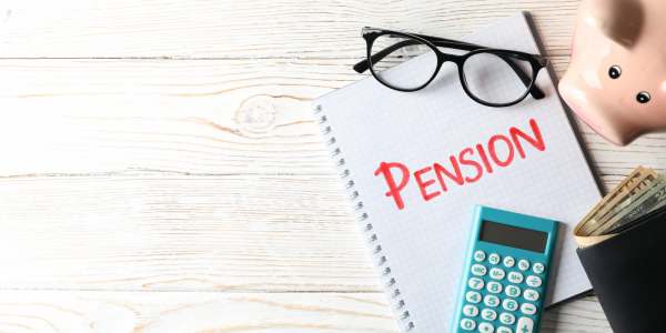 A desk with a piggy bank, a wallet,pair of glasses, a calculator and a notepad with the word 'PENSION' written on the front in red marker pen.