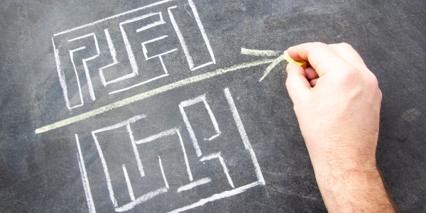 two hand drawn mazes on a chalkboard, and a person drawing a straight arrow through the gap between the two. 